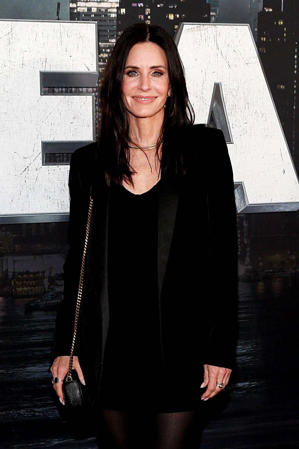Courteney standing before a "Z" sign, wearing a black blazer over a sheer top, smiling