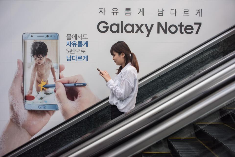 Una mujer mira a su teléfono móvil mientras desciende por una escalera mecánica, junto a un anuncio del Galaxy Note 7 de Samsung, el 11 de octubre de 2016 en Seúl (AFP | Ed Jones)