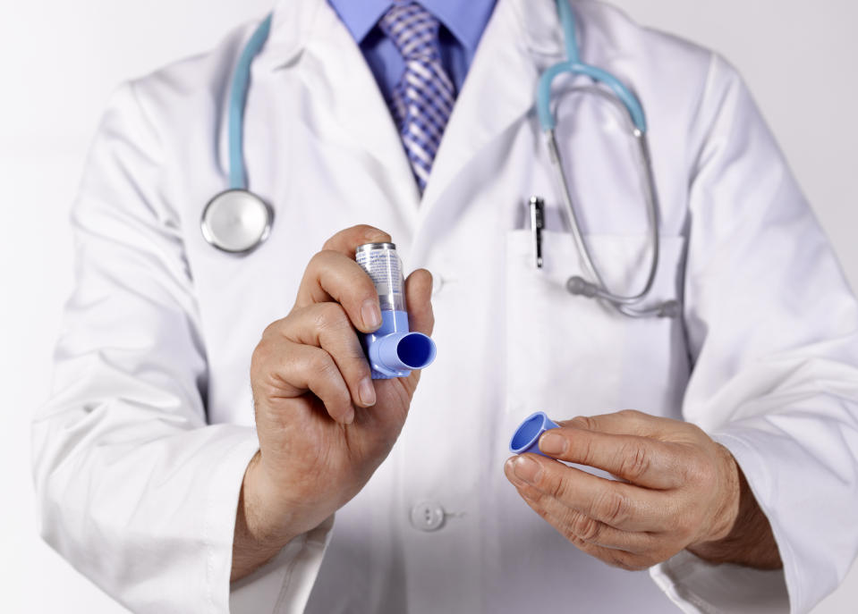 A person in a white lab coat and stethoscope holds an asthma inhaler, removing its cap, emphasizing medical care