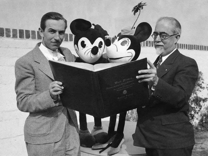 Black and white photo of Walt holding a large book with two original doll designs of Mickey and Minnie Mouse next to another man.