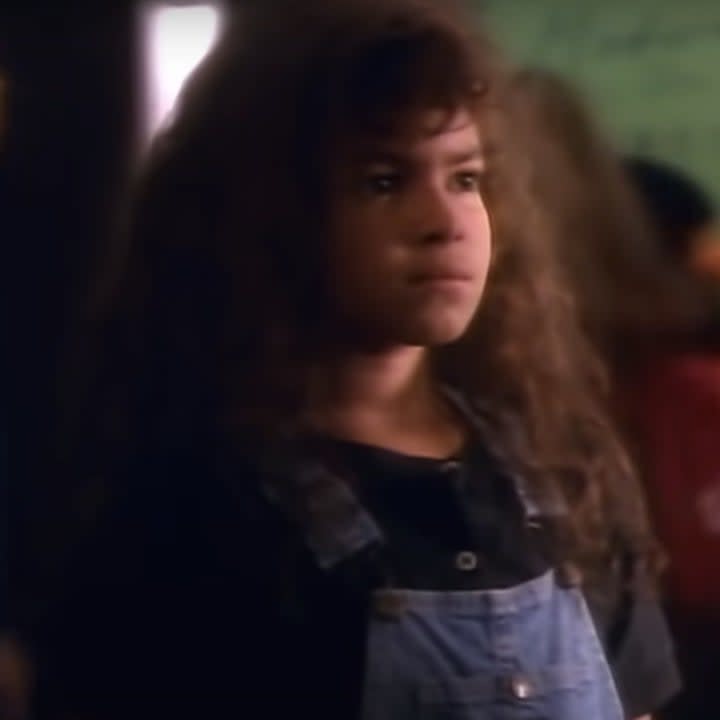 A young actress with brown curly hair wears denim overalls and a black t-shirt