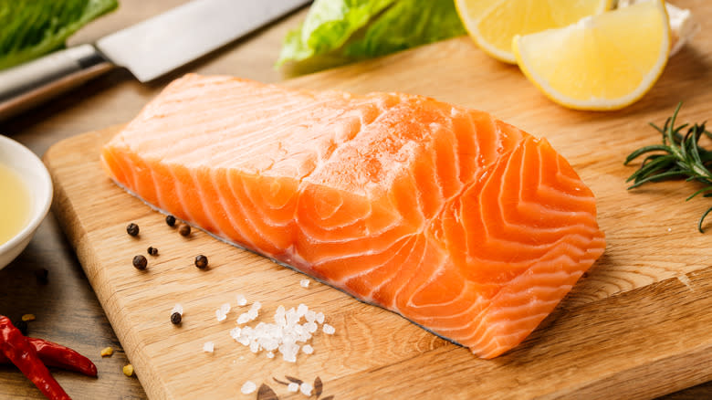raw salmon fillet on cutting board, knife, seasonings, lemon wedges