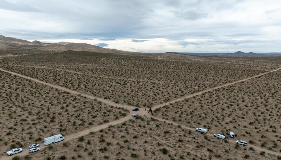 An aerial shot of two vehicles and several officials at a crossroads in the desert, with several white vehicles parked nearby