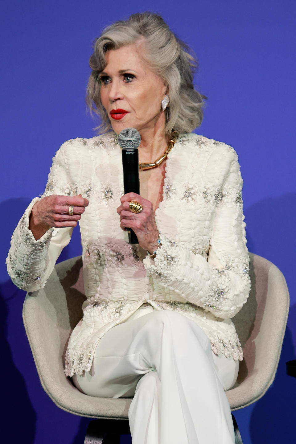 Jane Fonda wearing a textured, embellished blazer and white pants while speaking at an event