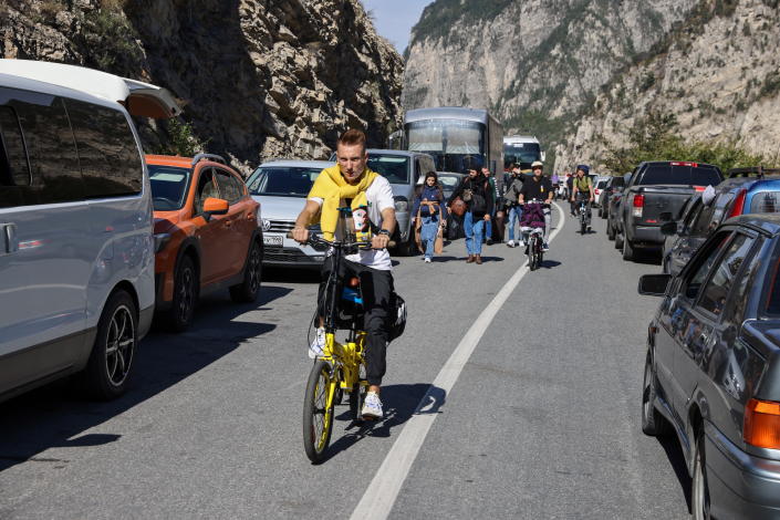 People walk and ride bicycles between rows of lined-up vehicles. 