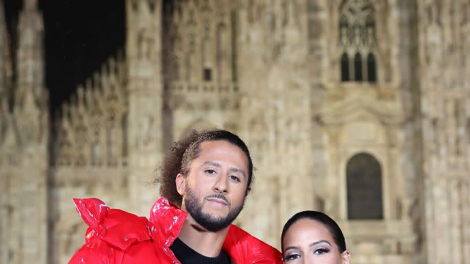 colin kaepernick and nessa diab pose for a photo while standing outside a cathedral, he wears a black shirt with red detailing and a red puffy coat, she wears a black turtleneck with a black puffy coat