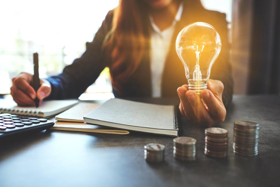 A person sitting at a table holding a lightbulb while writing on a notebook with stacks of coins on the table.