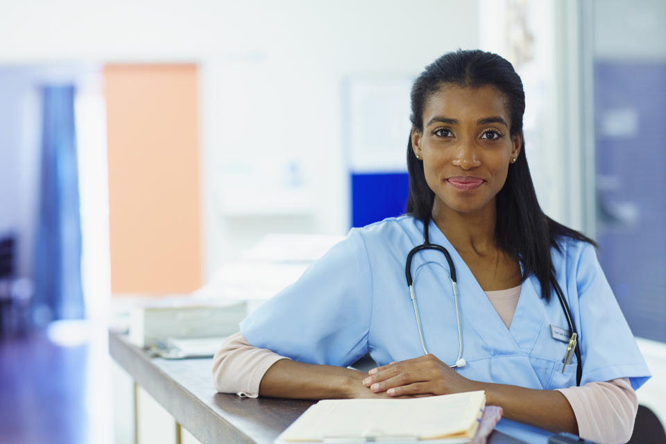 A woman in scrubs with a stethoscope around her neck smiles at the camera while standing at a desk in a medical setting