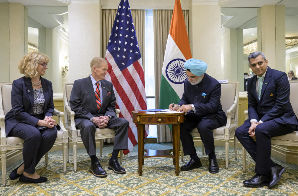 Four people sit facing forward and not towards each other.  They all wear suits.  One person wearing a light blue turban signs a document on a table in front of him.  Behind the middle two people are two flags.