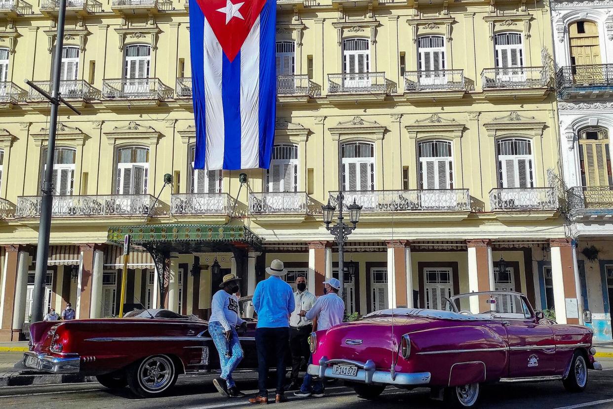 The drivers of old American cars talk near a Cuban flag outside the Inglaterra Hotel in Havana