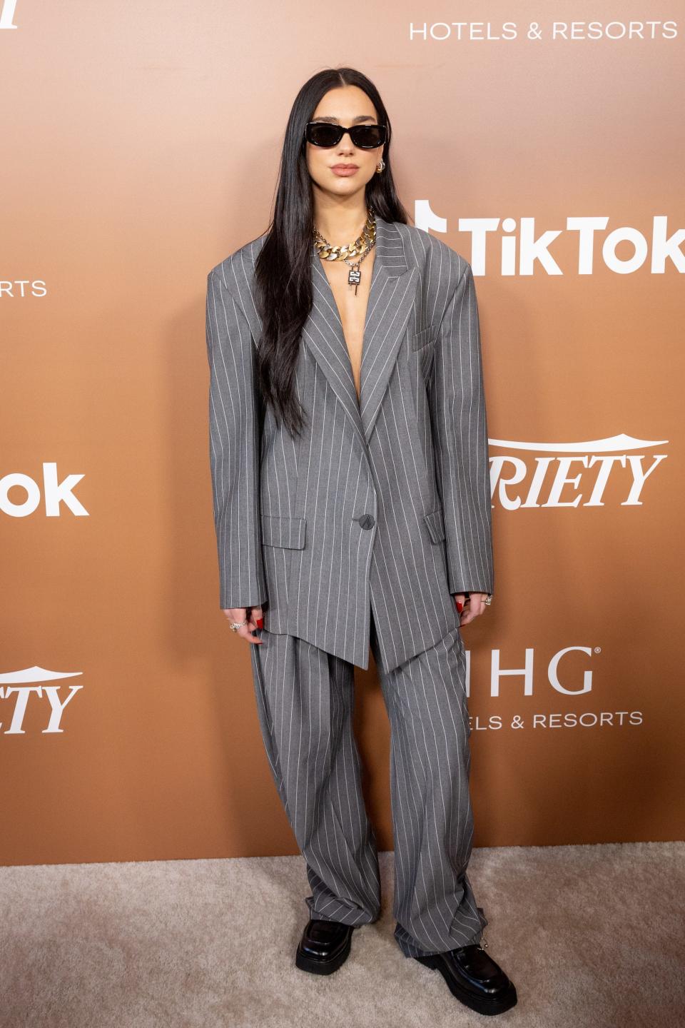 Dua Lipa stands in an oversized suit on a grey carpet.