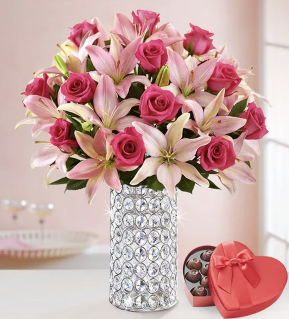 Pink lilies and roses in a crystal vase with heart shaped box of chocolates nearby. 