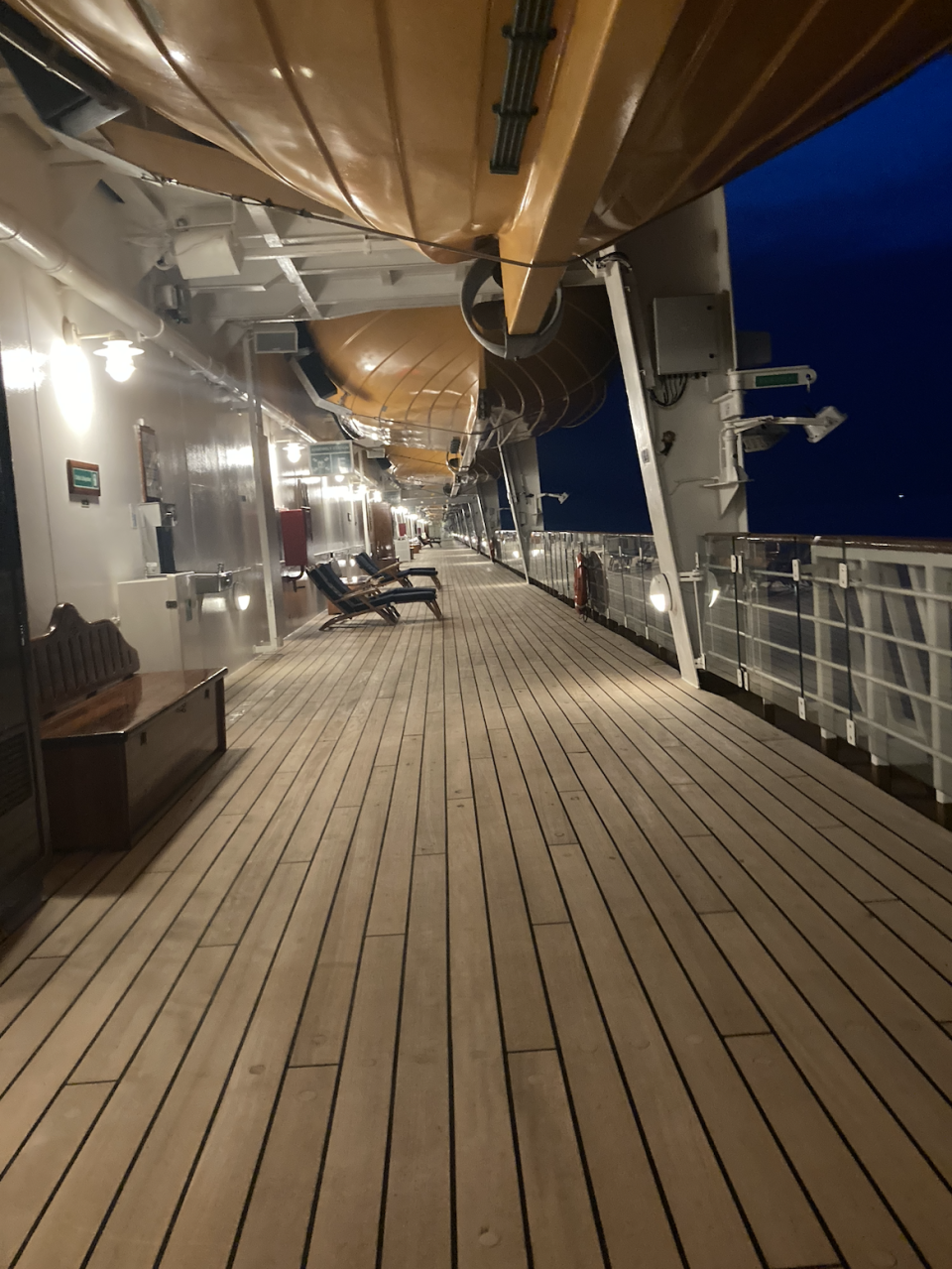 A ship's empty deck at night with wooden planks, lounge chairs, and lifeboats overhead