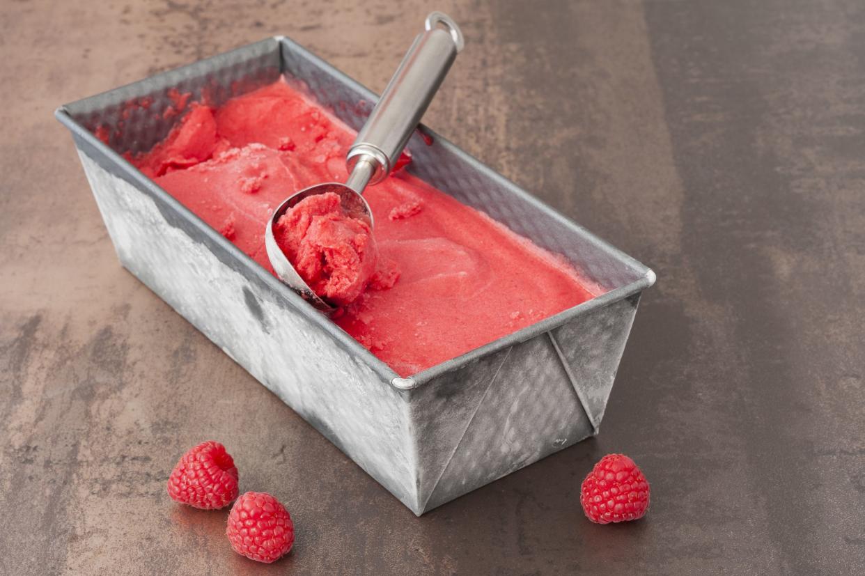 Fresh raspberry sorbet ice cream in a baking tin with a serving scoop on top. In the front are fresh raspberries