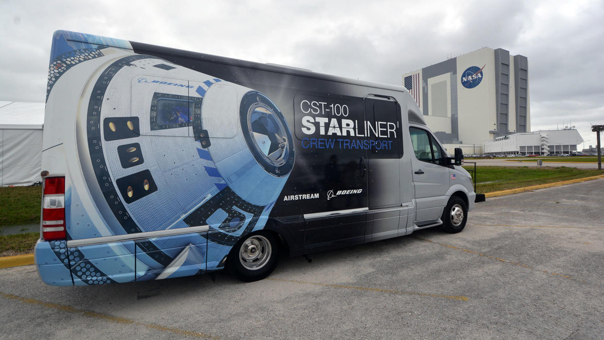 A large van with a white space capsule and the words "cst-100 starliner" painted on its side rolls down a road toward a building with the nasa logo on it. 