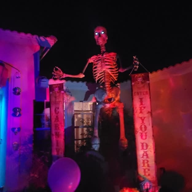 A skeleton at the entrance to a "carnival," with signs, at night