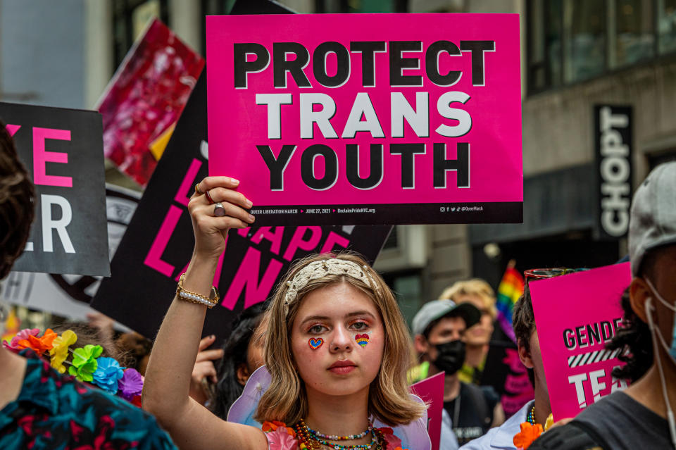 A young protester wearing heart stickers under each eye holds up a pink placard saying: Protect Trans Youth.