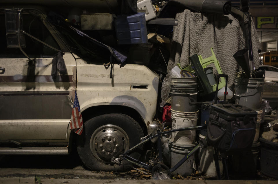 A tattered American flag hangs from the side of a van cluttered with personal belongings in a homeless RV encampment in downtown Los Angeles, Tuesday, Nov. 21, 2023. There is no single reason why Los Angeles became a magnet for homelessness. Two contributing factors: Soaring housing prices and rents punish those with marginal incomes, and a long string of court decisions made it difficult for officials to clear encampments or relocate homeless people from parks and other public spaces. (AP Photo/Jae C. Hong)