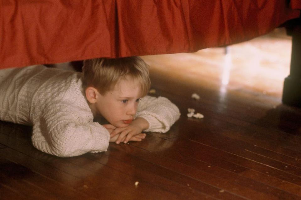 Kevin McCallister wears a white cable-knit sweater and lies on his stomach under a bed with a red bedskirt in a still from the original "Home Alone" movie.