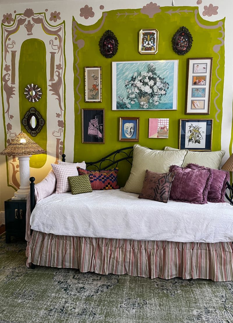 Skirted daybed with purple and green throw pillows in front of green wall with decorative paint featuring hung artwork.