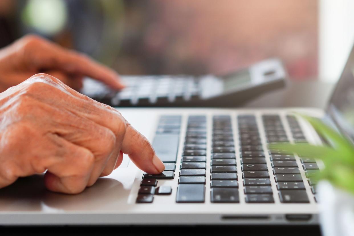 Side-view closeup of senior woman's hand on a MacBook while the other hand is using a calculator, blurred background