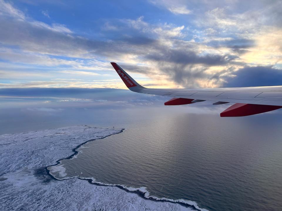A wing of a plane flying over a snowy area with a large body of water and ice along the edges. A sunrise with a yellow and blue sky sits above the water
