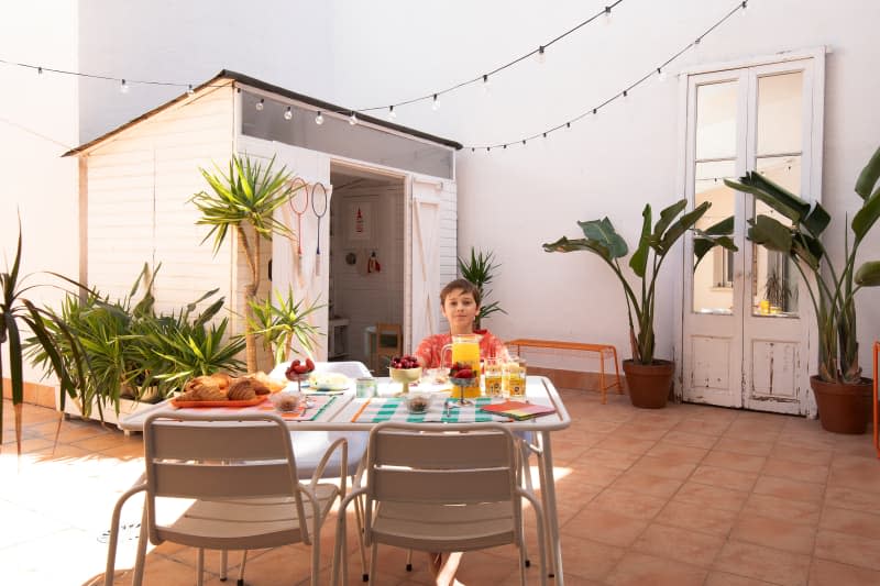 Child sitting at outdoor table on tile patio with small playroom and string lights