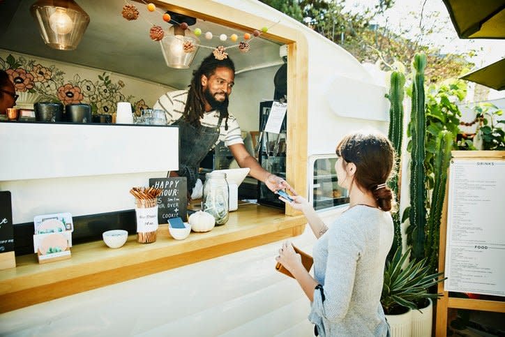 A woman handing her credit card through the window of a food truck to the owner standing inside wearing an apron.