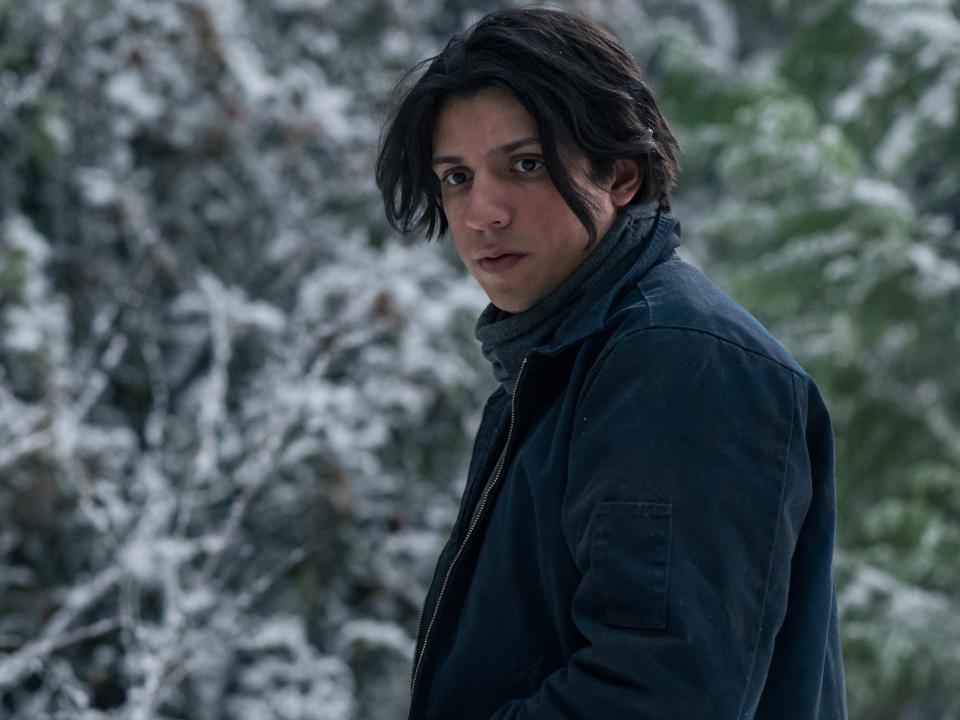 Teen Travis stands in the snow wearing blue jeans, a navy jacket, and a scarf.