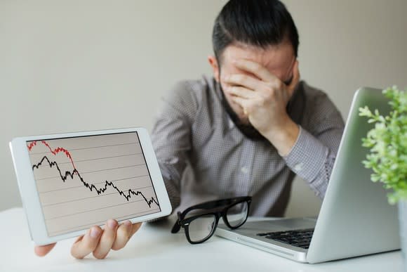 A frustrated investor with his hand covering his face holding up a tablet with a plunging stock chart.