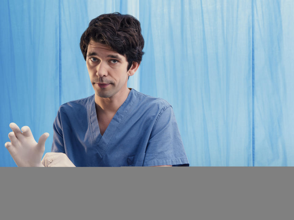 Ben Whishaw as Adam Kay in This Is Going To Hurt,  standing in front of a blue hospital curtain wearing blue surgical scrubs and pulling on a pair of rubber surgical gloves