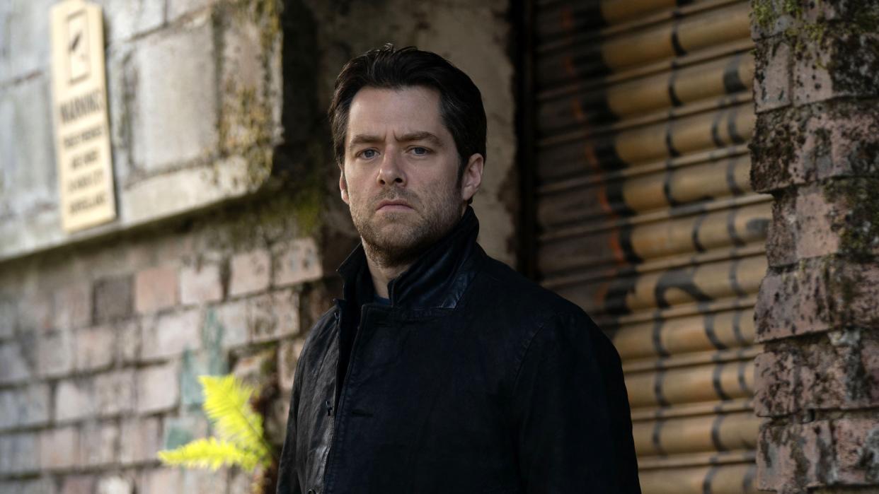  Richard Rankin in a dark jacket as Rebus stands in an alleyway in Rebus. 