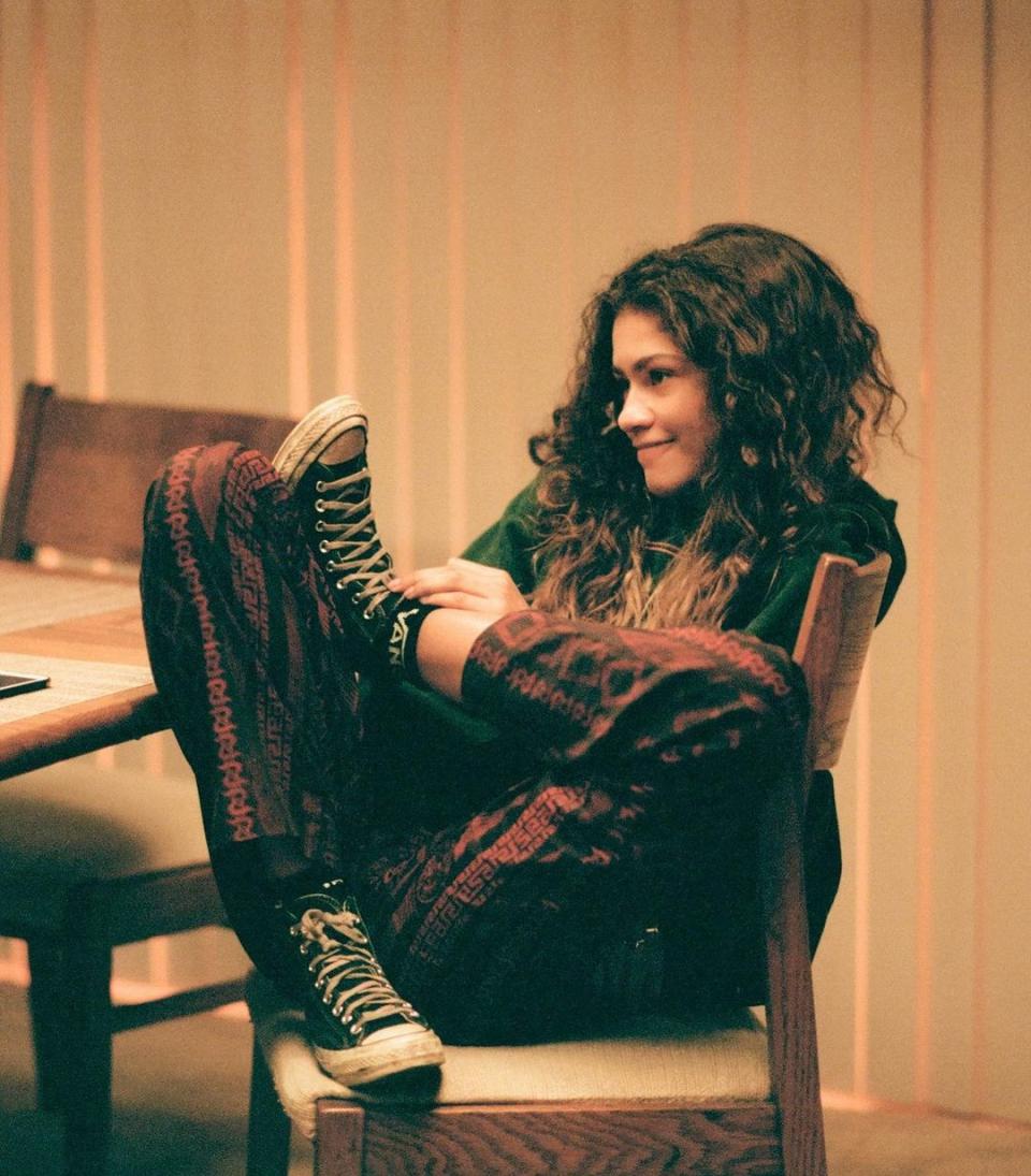 Euphoria character Rue Bennett sits curled up on a dining chair wearing skater clothes and sneakers with her dark curly hair worn loose.