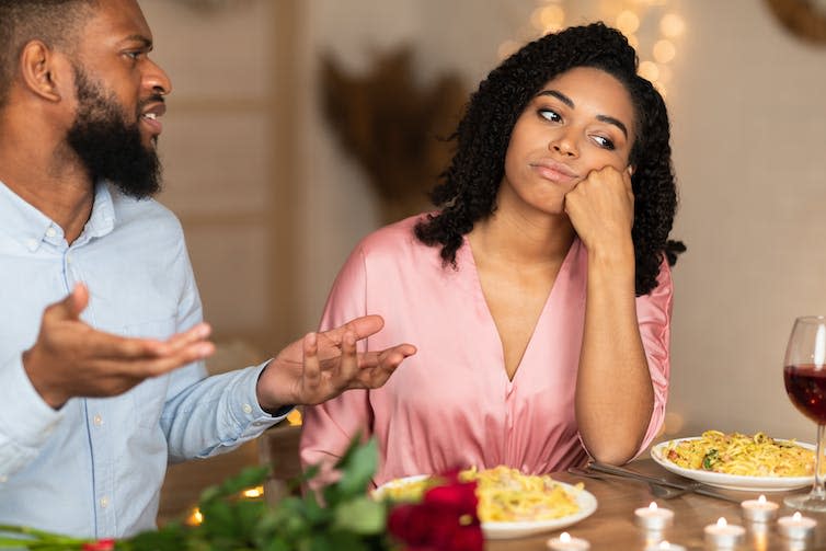 A young woman seated at a dinner table looks unhappy, resting her head on her fist, as the man seated next to her speaks while gesturing with both of his hands