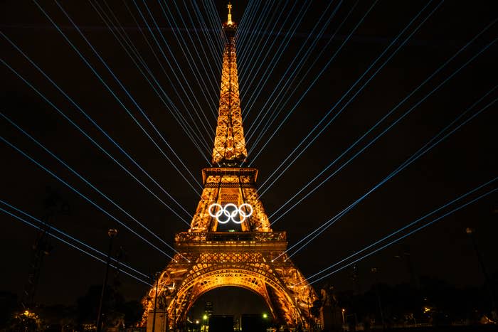 The Eiffel Tower lit up at night with the Olympic rings displayed and beams of light radiating from it, celebrating the upcoming Olympic Games