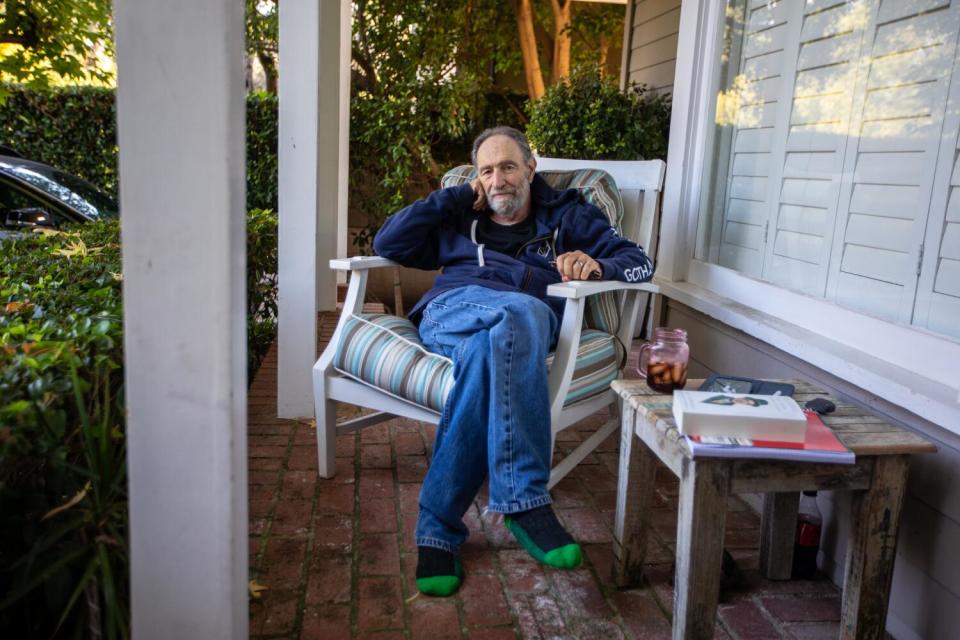 An older man sits with his legs crossed in a chair on a porch.