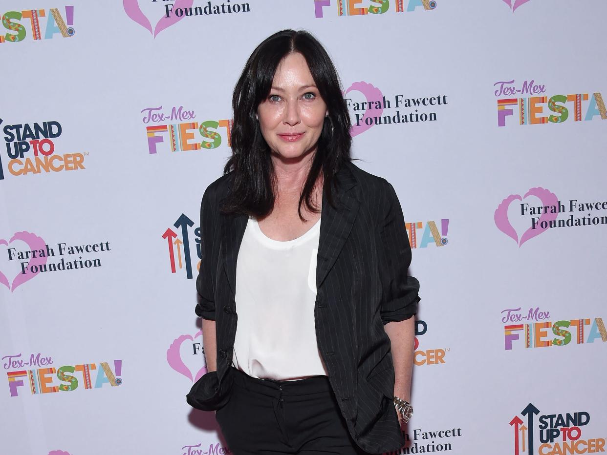 Shannen Doherty in a black blazer and white top standing on a red carpet.