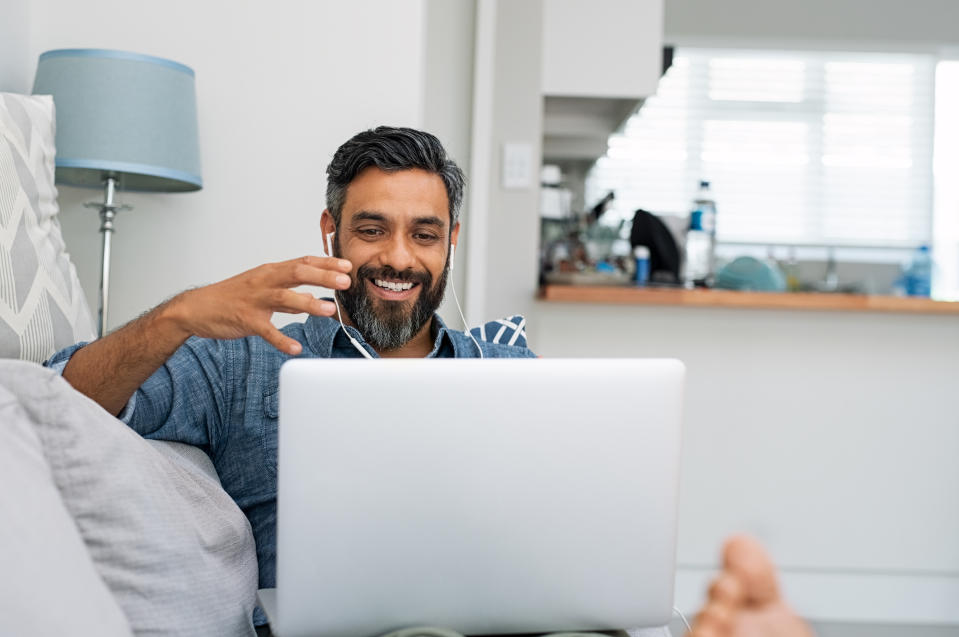 Happy mature man relaxing on couch while video calling using laptop at home. Latin man sitting on sofa and making a video call. Smiling middle eastern businessman doing online video chat while gesturing with hands.