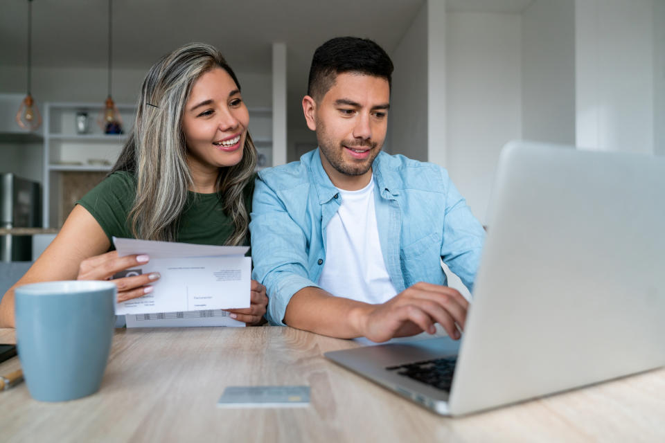 Happy latino couple paying bills online with laptop at home and smiling - financial technology concept