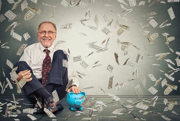 Older man sitting next to a blue piggy bank with money raining down around him.