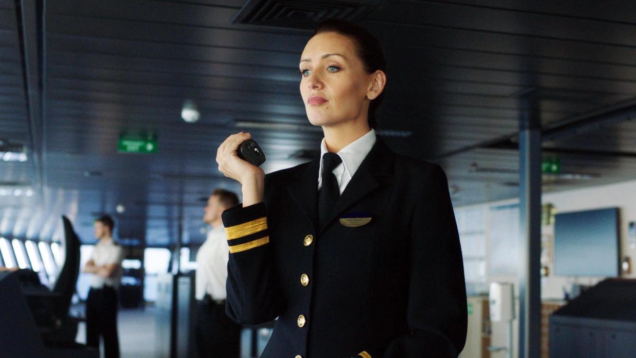  First Officer Kate Woods (Catherine Tyldesley) stands on the bridge of the cruise ship, in full uniform with blazer, holding a radio that she's speaking into. 