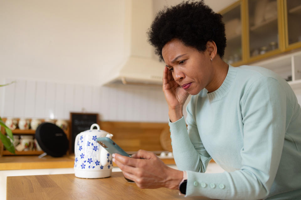 A woman in a kitchen looks concerned while holding a smartphone. She leans on a wooden counter with a kettle in the background