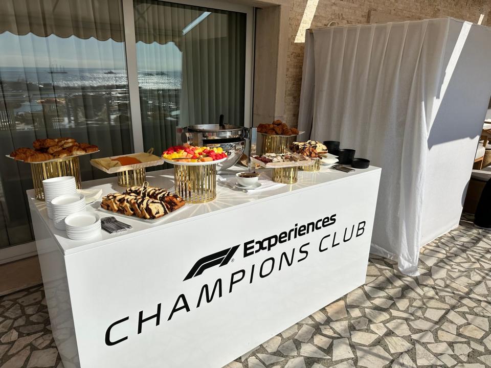 Ein weißer Tisch, gefüllt mit Gebäck, Obst und anderen Lebensmitteln. Auf dem Tisch steht in schwarzer Schrift "Experiences Champions Club".