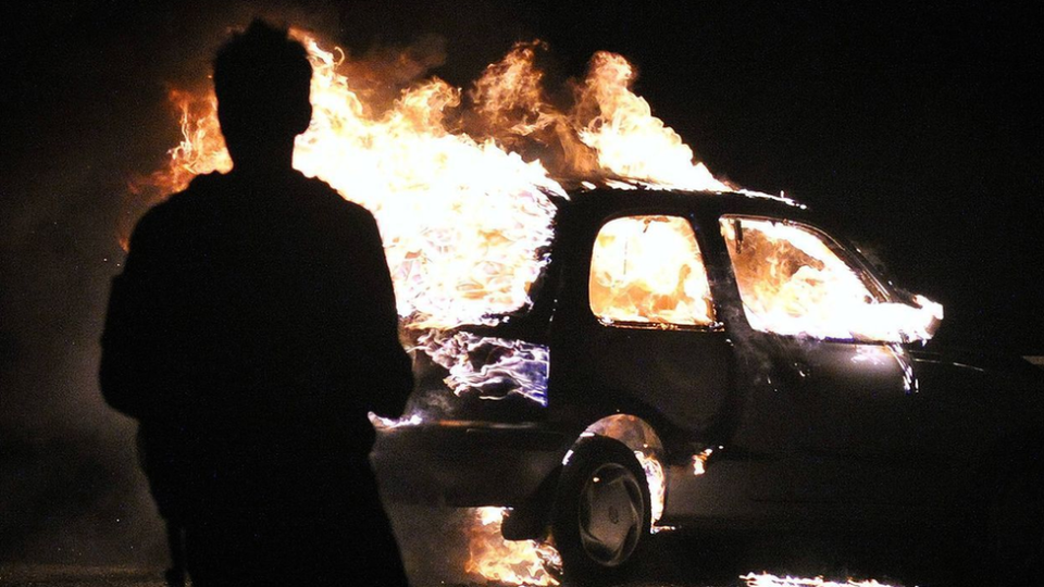 A person stands in front of a burning car at night