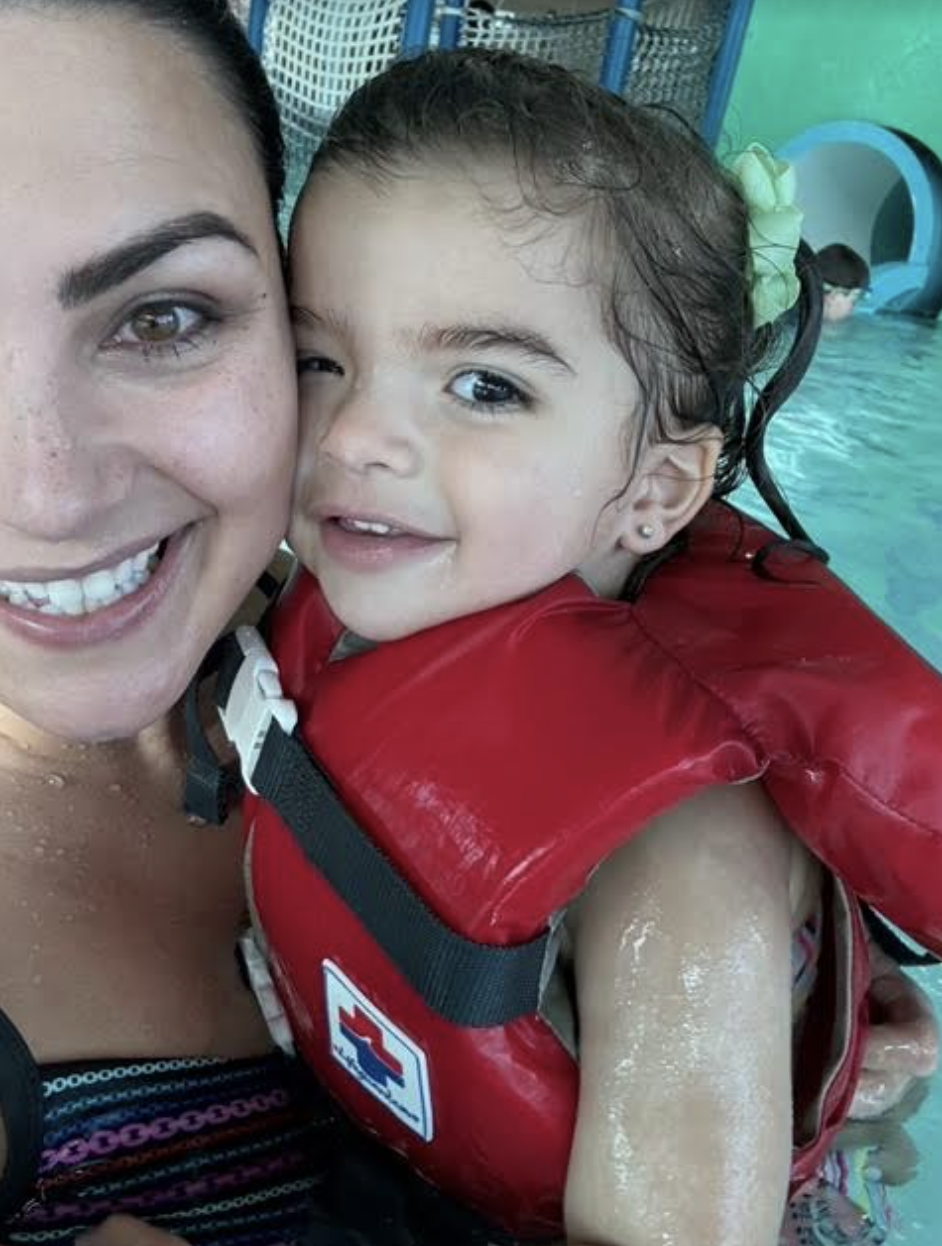 A woman smiles at the camera while holding a young child wearing a life vest. Both are in a pool. Names are not provided