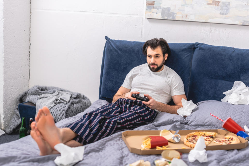 A man playing video games as food an trash litter his bed