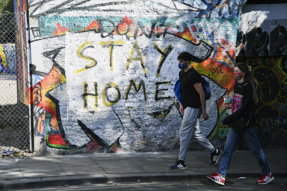 A couple wearing protective masks to protect against coronavirus walk past a mural reading 'Stay home', in the Shoreditch area, as the lockdown continues due to the coronavirus outbreak, in London, Saturday, April 25, 2020. (AP Photo/Alberto Pezzali)