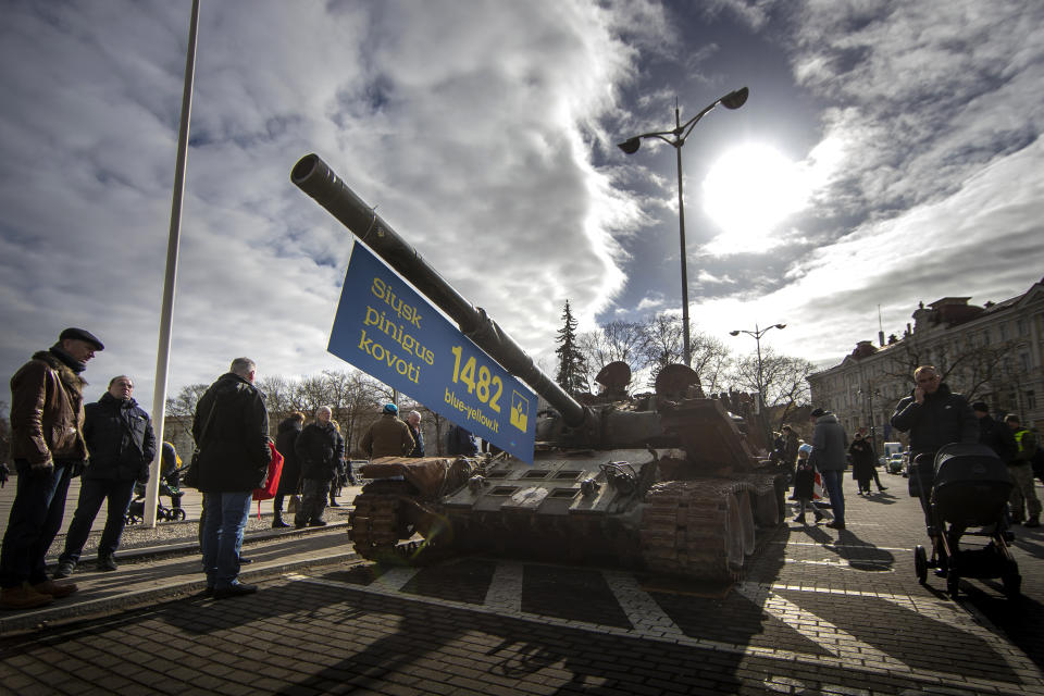 People stand by a destroyed Russian tank T-72B, installed as a symbol of war marking the first anniversary of Russia's full-scale invasion of Ukraine, and decorated with a banner with writing reading "Send money to fight", at Cathedral Square in Vilnius, Lithuania, Wednesday, March 1, 2023. Some ethnic Russians in the Baltic states have placed flowers at displays of burnt-out Russian tanks seized by Ukrainians, making a gesture of homage and support for Russia's war against Ukraine. (AP Photo/Mindaugas Kulbis)