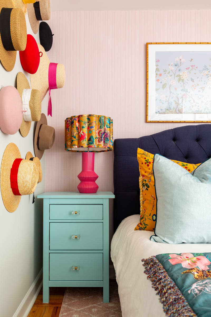 A pink lamp with a floral shade next to a sunhat hung on a wall.