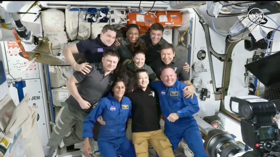 Nine astronauts float in a mostly white module of a space station, all smiling for photos.